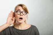 A surprised looking woman wearing glasses holds her hand up to her ear