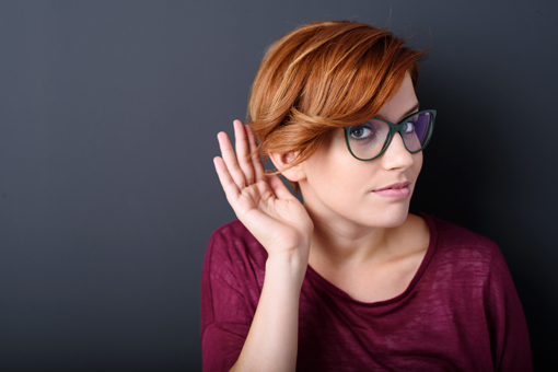 A woman with red hair, holds up her hand to her ear, as to gesture listening