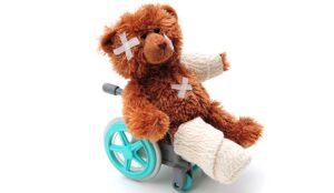 A teddy bear sits in a little wheelchair, with a leg in a bandage and plasters on its head