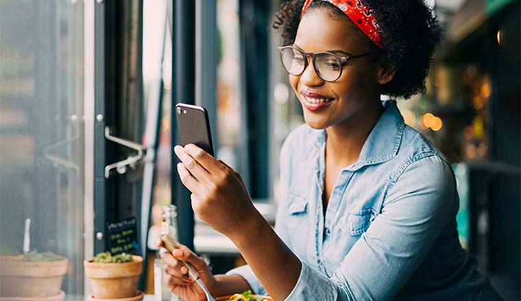 woman smiling and reading texts on a cellphone while sitting alone at a counter in a cafe enjoying a meal