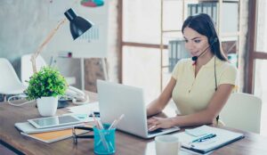 Smiling businesswoman is using headset when talking to customer and typing on her laptop