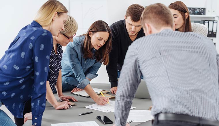 Motivated group of young business people grouped around a table listening to a female team leader or manageress explain paperworkMotivated group of young business people grouped around a table listening to a female team leader or manageress explain paperwork