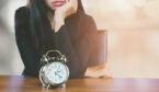 A clock is sat on a desk in front of a bored looking lady