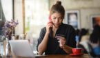 A woman is looking at her credit card while on her phone, sat at a desk next to a computer