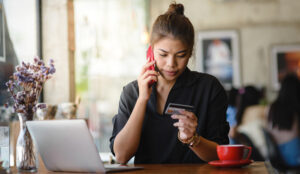 A woman is looking at her credit card while on her phone, sat at a desk next to a computer