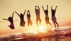 Big group of happy friends having fun and jumps in water against sunset