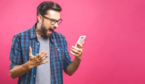 Young caucasian man angry, frustrated and furious with his phone, angry with customer service over pink background.