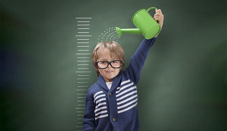 Young boy trying to make himself taller with watering can measuring his growth in height against a blackboard scale