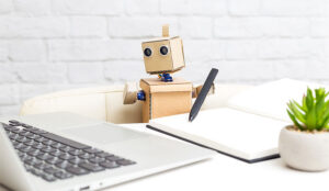 A photo of a cardboard robot holding a pen and looking at a laptop