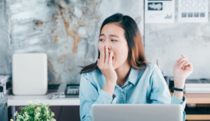 A bored looking woman holds her hand in front of her face, while working at a laptop