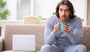 A person drinking tea whilst sat on a sofa next to a laptop