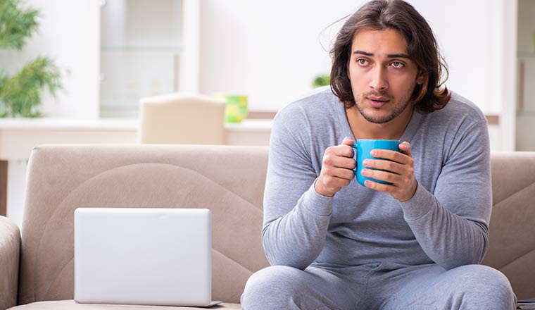 A person drinking tea whilst sat on a sofa next to a laptop