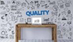 A picture of the word quality doodles on a wall behind a desk with laptop