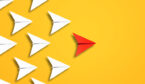 A red paper aeroplane leading a group of white ones on a yellow background