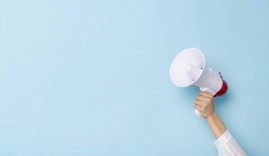 Hand holds a megaphone from a hole in the wall on blue background