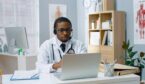 A physician in white coat speaking on video call in headset on laptop