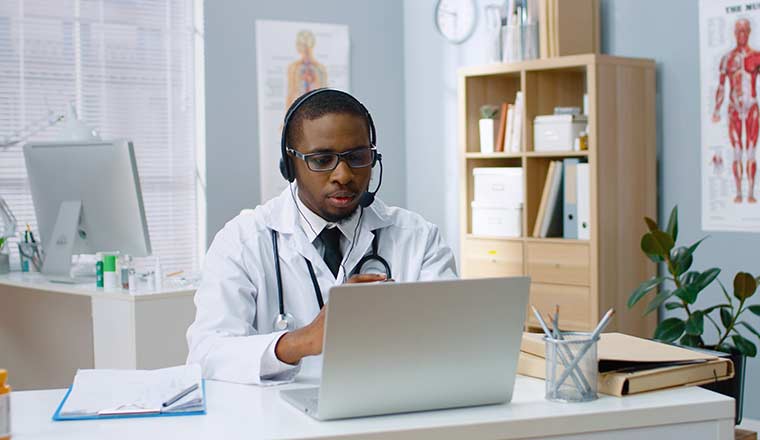 A physician in white coat speaking on video call in headset on laptop