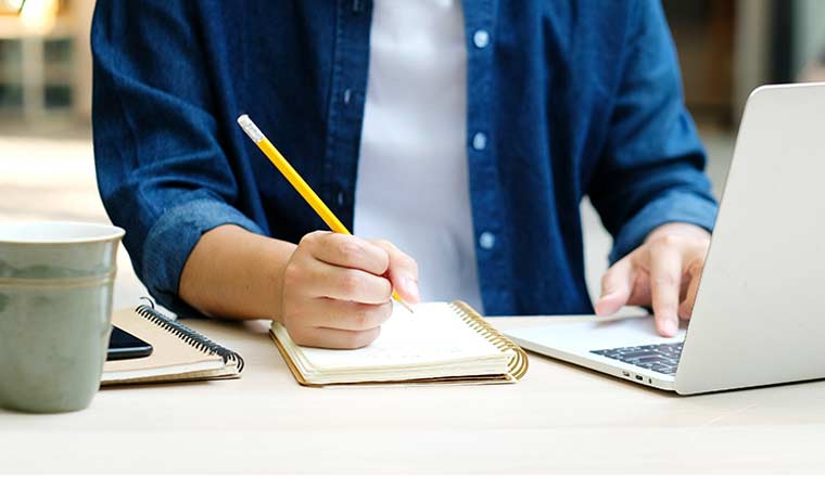 Person hand writing on notebook while using laptop computer