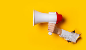 Hand holds a megaphone from a hole in the wall on a yellow background