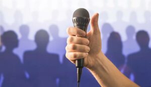 Hand holding microphone and showing thumbs up in front of a crowd of silhouette people