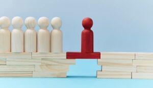 Wooden figures in line, one red person miniature ahead on bridge gap