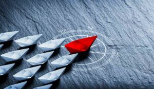 Red Paper Boat On Compass Leading A Fleet Of Small White Boats