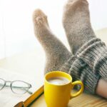 Person relaxing on a day off with legs in warm socks on table with morning coffee and reading book
