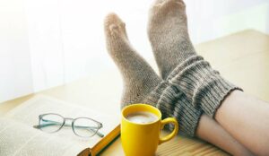 Person relaxing on a day off with legs in warm socks on table with morning coffee and reading book