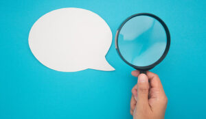 Top view of a blank white speech bubble and hand holding a magnifying glass on a blue background.