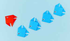 Red origami paper fish changing direction while swimming ahead of line of blue fish.