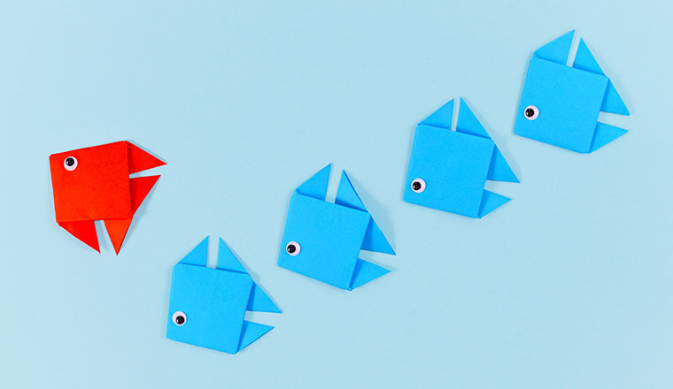 Red origami paper fish changing direction while swimming ahead of line of blue fish.