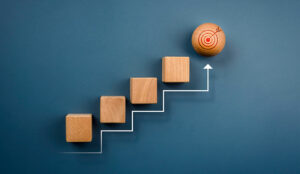 White rising arrow with wooden cube blocks arranged as a chart stair steps up to wood sphere with target icon on top