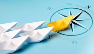 A yellow paper boat on a compass leading a trail of white paper boats