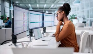 Person sat at desk with three screens with spreadsheets